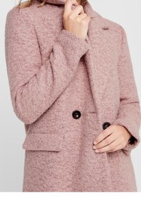 Rosa texturerad kappa med krage, dubbla svarta knappar och sidofickor. Tyget verkar ull-liknande med en mjuk, något ludden yta.