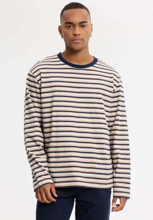 Hombre que lleva una camisa beige de manga larga con rayas horizontales azul marino y rojas y pantalones azul marino, de pie frente a un fondo liso.