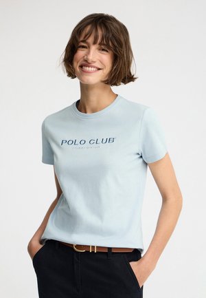 Lächelnde Frau mit kurzen braunen Haaren, die ein hellblaues "Polo Club"-T-Shirt trägt, das in schwarze Hose mit braunem Gürtel gesteckt ist, Hände in den Taschen.