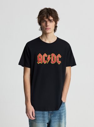 Giovane uomo con capelli scompigliati che indossa una maglietta nera con il logo AC/DC rosso e giallo e jeans blu sbiaditi, in piedi davanti a uno sfondo chiaro e semplice.