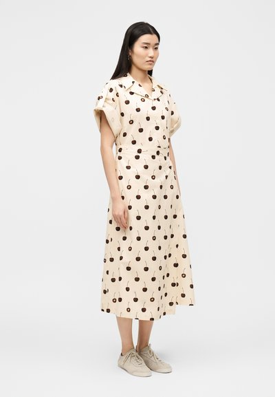 Γυναίκα που στέκεται, φορώντας ένα μίντι φόρεμα σε κρεμ απόχρωση με σχέδιο κεράσια, κοντά μανίκια, γιακά και μπεζ αθλητικά παπούτσια, στραμμένη ελαφρώς προς τα δεξιά.