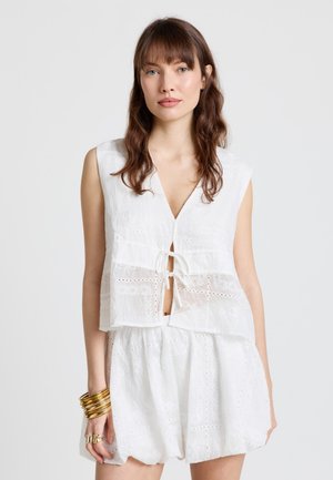 Jeune femme aux longs cheveux bruns portant un haut sans manches blanc en broderie anglaise avec des liens à l'avant et une jupe blanche assortie, accessoirisée de bracelets en or.