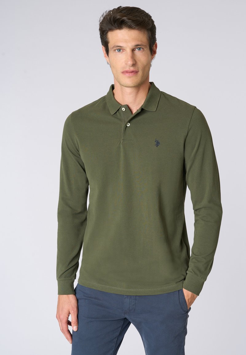 Polo a maniche lunghe di colore verde oliva con tre bottoni, tessuto testurizzato e un piccolo logo navy sul lato sinistro del petto, abbinato a pantaloni blu.