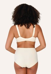 Grudnjak boje kreme s širokim naramenicama i podesivim kopčama, upareni s gaćicama visokog struka boje kreme. Glatka tekstura, minimalistički dizajn.