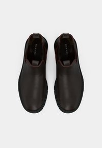 Une paire de bottines slip-on en cuir marron foncé avec des semelles noires, vue de dessus sur un fond blanc.