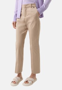 Beige tailored pants with a smooth texture, featuring a flat front and side pockets, paired with light beige sandals with logo accents.