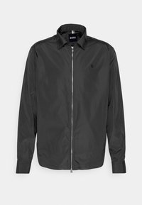 Sort letvægtsjakke med lynlås, krave, lange ærmer og subtilt logo på venstre bryst, vist på en ensfarvet baggrund.