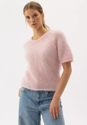 Jonge vrouw die een pluizige roze korte mouwen trui en lichtblauwe spijkerbroek draagt, staand tegen een effen witte achtergrond.