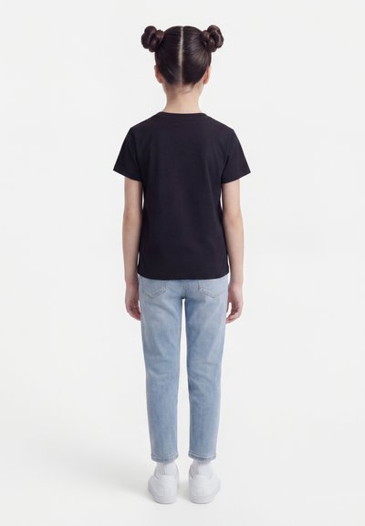 Παιδί που στέκεται με την πλάτη, φοράει μαύρο μπλουζάκι, ανοιχτό μπλε τζιν, λευκές κάλτσες, λευκά αθλητικά παπούτσια και έχει τα μαλλιά του πιασμένα σε δύο κότσους.