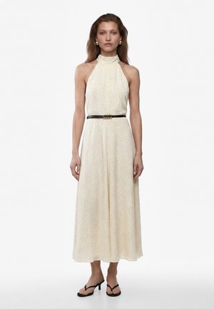 Donna in piedi che indossa un vestito crema senza maniche con collo alto e piccoli pois, cintura nera, orecchini dorati e sandali neri aperti con tacco.