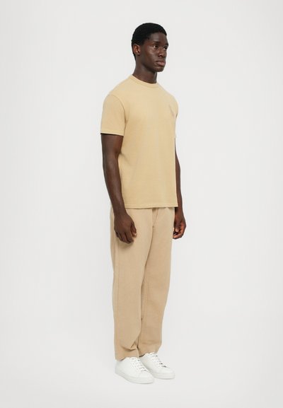 Beige bomull t-shirt och matchande lösa byxor, båda med en mjuk textur. Vita sneakers med enkel design utan synliga logotyper eller mönster.