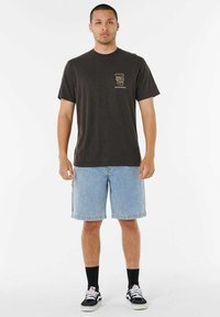 Donkergrijze katoenen t-shirt met een klein gouden logo op de borst, gecombineerd met lichtblauwe denimshorts. Zwarte sokken en zwarte sneakers maken de outfit compleet.
