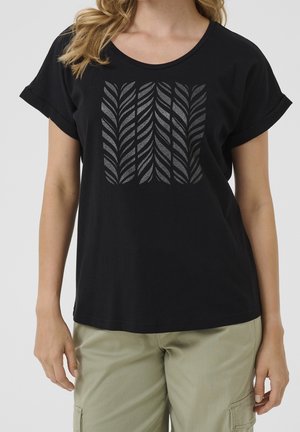 Kvinde iført en sort T-shirt med korte ærmer og optrukne manchetter samt et gråt abstrakt bladmotiv på brystet, kombineret med lyse khakibukser.