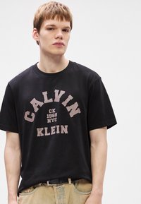 Črna bombažna majica s kratkimi rokavi, ki ima na sprednji strani velik bel napis "CALVIN KLEIN", pod njim pa v manjšem fondu "CK 1968 NYC". Ima okroglo ovratnico.