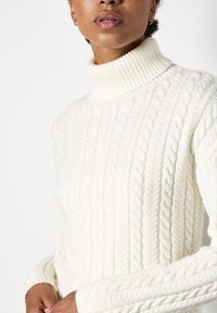 Maglione a collo alto a trecce color crema con colletto e polsini a costine. Presenta motivi testurizzati e un tessuto morbido e spesso.