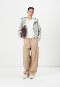 Sweat à capuche gris zippé, haut blanc à boutons, pantalon beige ample et baskets blanches. Portant un sac bordeaux. Fond uni.