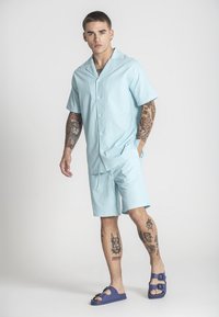 Camicetta azzurro chiaro a maniche corte e pantaloni abbinati realizzati in un tessuto texturizzato, completati da sandali blu. Presenta bottoni e una vestibilità rilassata.