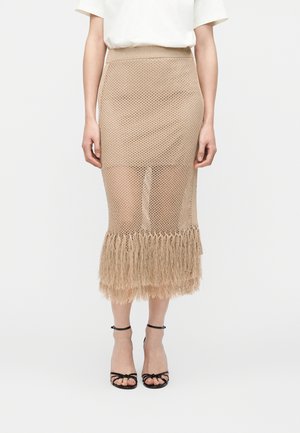 Béžová pletená midi sukňa s lemom s strapcami, nosená s čiernymi sandálmi s remienkami a bielym krátkym rukávom na jednofarebnom pozadí.