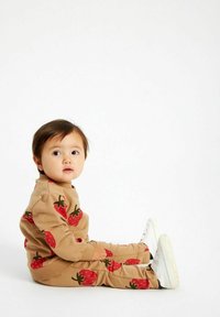 Set di felpa e pantaloni color marrone chiaro con stampe di fragole rosse, con polsini a costine. Il bambino è seduto con sneakers bianche su uno sfondo neutro.