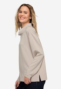 Sudadera beige con cuello de cowl, de manga larga, con aberturas laterales y un ajuste relajado, fabricada con material suave. Llevada con jeans oscuros.