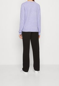 Persona de pie, de espaldas, vistiendo un suéter de manga larga lavanda, pantalones negros y zapatillas blancas, frente a un fondo liso.