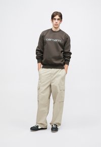 Hanorac maro cu logo alb, pantaloni cargo bej cu buzunare laterale și pantofi negri cu accente albe. Modelul stă cu mâinile în buzunare.