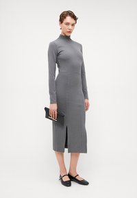 Grå langermet kjole med høy hals, midt på leggen lengde og splitt foran. Stylert med svarte flate sko og en matchende clutch. Myk stofftekstur.