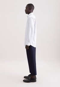 Camicia a righe bianche con maniche lunghe e colletto a punta, abbinata a pantaloni sartoriali blu navy e scarpe nere con lacci, vista di profilo.