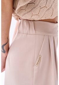 Beige plooienrok met een elastische tailleband, met een klein label op de zijkant met de tekst "blue shadow". Soepele textuur en op maat gemaakte pasvorm.