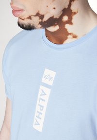 Světle modré bavlněné tričko se bílo vertikálním textem a logem "ALPHA". Design s kulatým výstřihem, hladká textura, krátké rukávy.