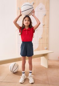 Rotes Poloshirt, marineblaue Shorts und weiße Sneaker mit schwarzen Streifen. Hält einen Basketball über ihrem Kopf, mit einer Kletterwand im Hintergrund.