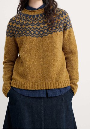 Personne portant un pull en maille jaune moutarde avec un motif géométrique bleu marine sur les épaules, superposé sur une chemise bleu marine et une jupe foncée.
