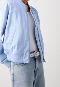 Jasnoniebieska kurtka bomber wykonana z gładkiego materiału, z ściągaczami na rękawach i przednim zamkiem błyskawicznym, noszona na dopasowanym białym topie, zestawiona z jasnymi dżinsami i paskiem.