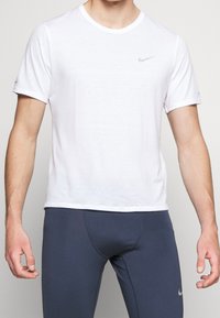 Homme portant un t-shirt de sport à manches courtes blanc et un legging de compression bleu marine, se tenant devant un fond clair.
