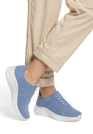 Hellblaue Stricksneaker mit weißen, dicken Sohlen, getragen zu beigen, hochgekrempelten Cordhosen vor weißem Hintergrund.