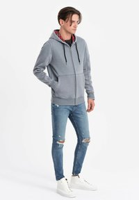 Felpa grigia con zip e rivestimento a quadri, abbinata a jeans blu strappati e sneakers alte bianche. Presenta lacci e tasche frontali.