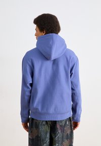 Purple hoodie with a large hood, ribbed hem, and contrast stitching. Worn over patterned, multi-colored pants with a foliage design.