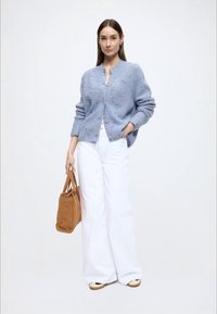 Svetlomodrý pletený sveter na gombíky vpredu, doplnený o biele široké nohavice. Modelka drží hnedú semišovú kabelku.