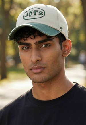 Junger Mann mit einer beige-grünen New York Jets Kappe und einem schwarzen Hemd, der im Freien vor unscharfem Baumbewuchs steht.