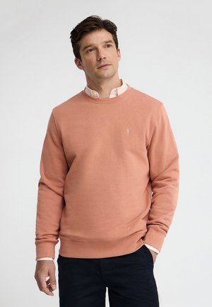 Man draagt een lichtroze crewneck trui over een lichtroze overhemd met kraag en zwarte broek, staand tegen een effen lichte achtergrond.