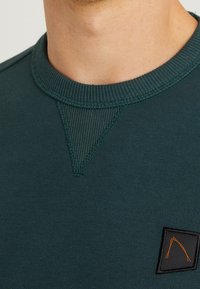 Detailní záběr na tmavě zelenou mikinu s kulatým výstřihem, která má žebrované V-šití a malou černou nášivku s oranžovým vyšitým vzorem.