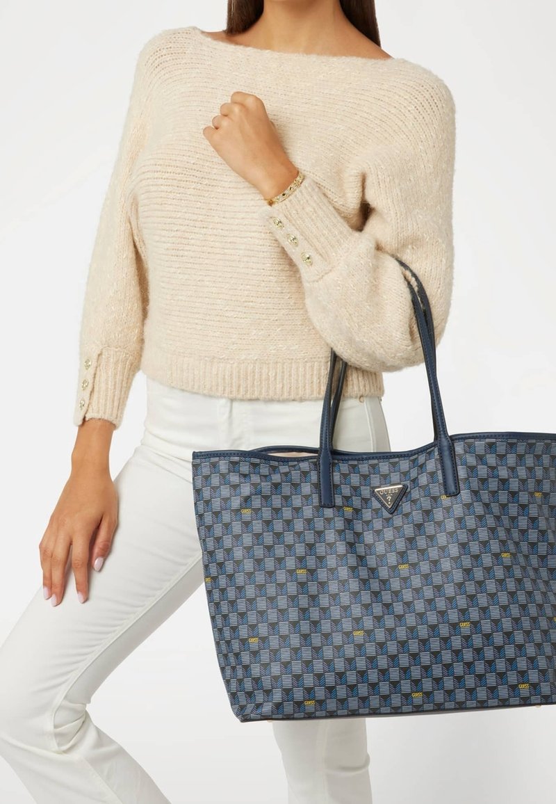 Femme portant un pull en maille beige et un pantalon blanc tient un sac cabas bleu à motifs Guess avec des anses foncées sur son bras.