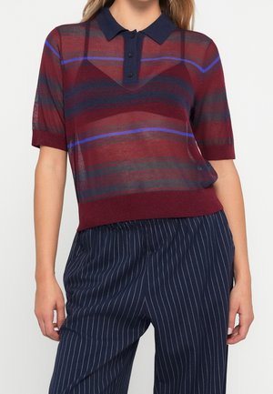 Vrouw draagt een doorschijnend, kort mouw polohemd met horizontale rode, marineblauwe en blauwe strepen, gecombineerd met marineblauwe, fijn gestreepte broek.