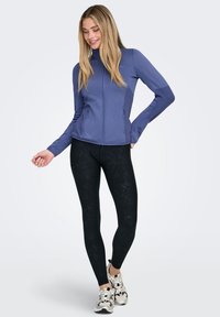 Lightweight, fitted purple zip-up jacket with high collar, textured side panels, and black patterned leggings with a smooth finish.