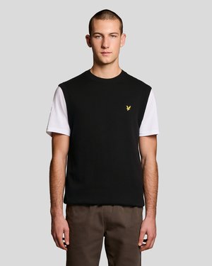 Fiatal férfi rövid hajjal, fekete ujjatlan pulóvert visel fehér rövid ujjú ing fölött és barna nadrágot, világos háttér előtt áll.