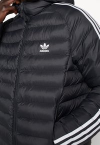 Svart vadderad jacka med dragkedjestängning, försedd med huva, vit Adidas-logotyp och tre vita ränder längs ärmarna. Slät textur.