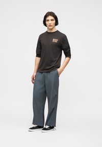 Νέος άνδρας με σκούρα μαλλιά, φορέας μαύρου μακρυμάνικου μπλουζάκι με σχέδιο, φαρδιά γκρι παντελόνια και μαύρα αθλητικά παπούτσια, στέκεται με το ένα χέρι στην τσέπη.