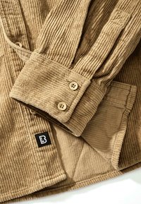Chemise en velours côtelé marron clair avec poignets boutonnés et une petite étiquette logo noire et blanche sur l'ourlet.