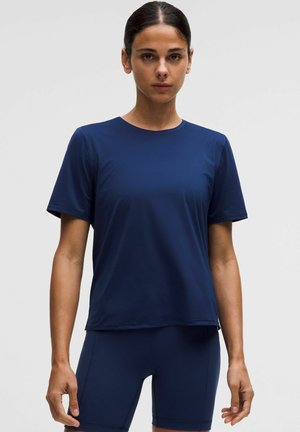 Frau mit zurückgebundenem dunklem Haar, die ein marineblaues Kurzarmshirt und passende marineblaue Radlerhosen vor einfachem Hintergrund trägt.