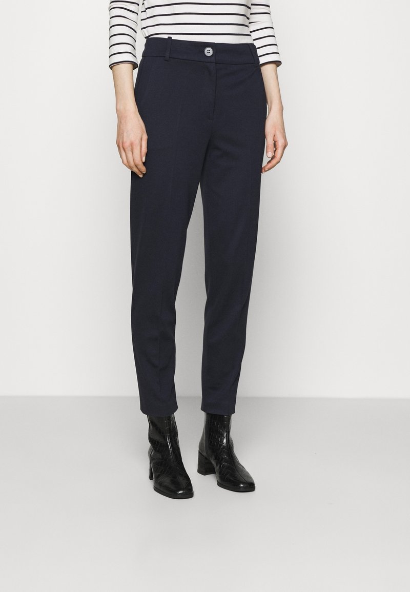 Vrouw in marineblauwe op maat gemaakte broek, zwarte enkellaarsjes met hakken en een witte blouse met lange mouwen en zwarte horizontale strepen, staande op een grijze vloer.